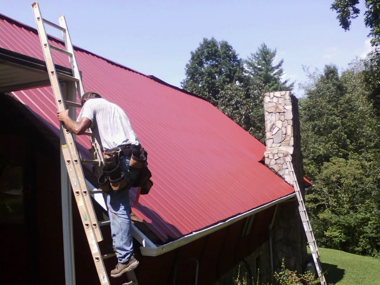 Roofer on a ladder beside a red metal roof and stone chimney — Soffit & Fascia Repair by Roofing Brothers in Milwaukee